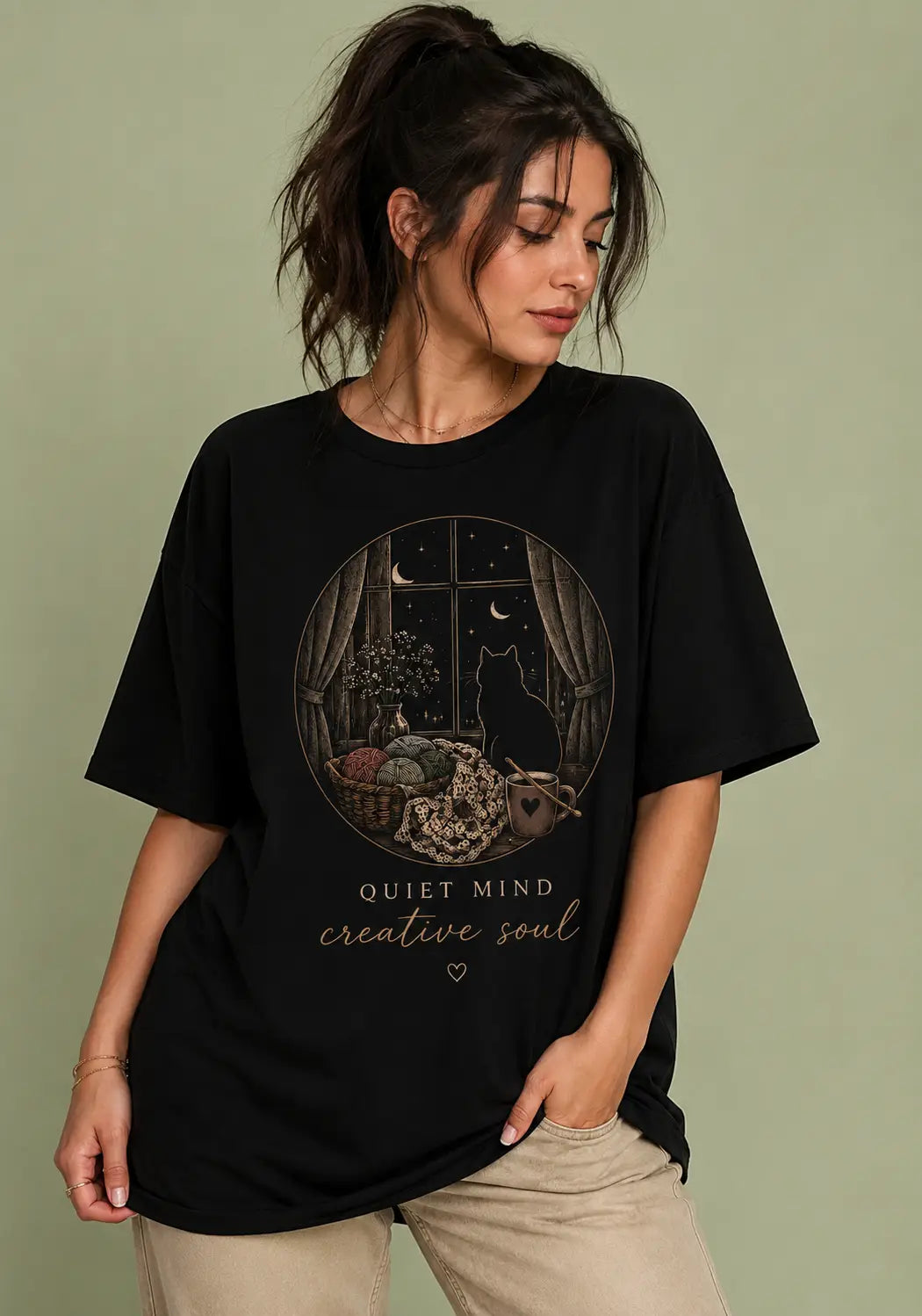 A woman wearing an oversized black graphic t-shirt for crochet artists with a circular illustration of a cat by a window at night and the text 'QUIET MIND creative soul'.