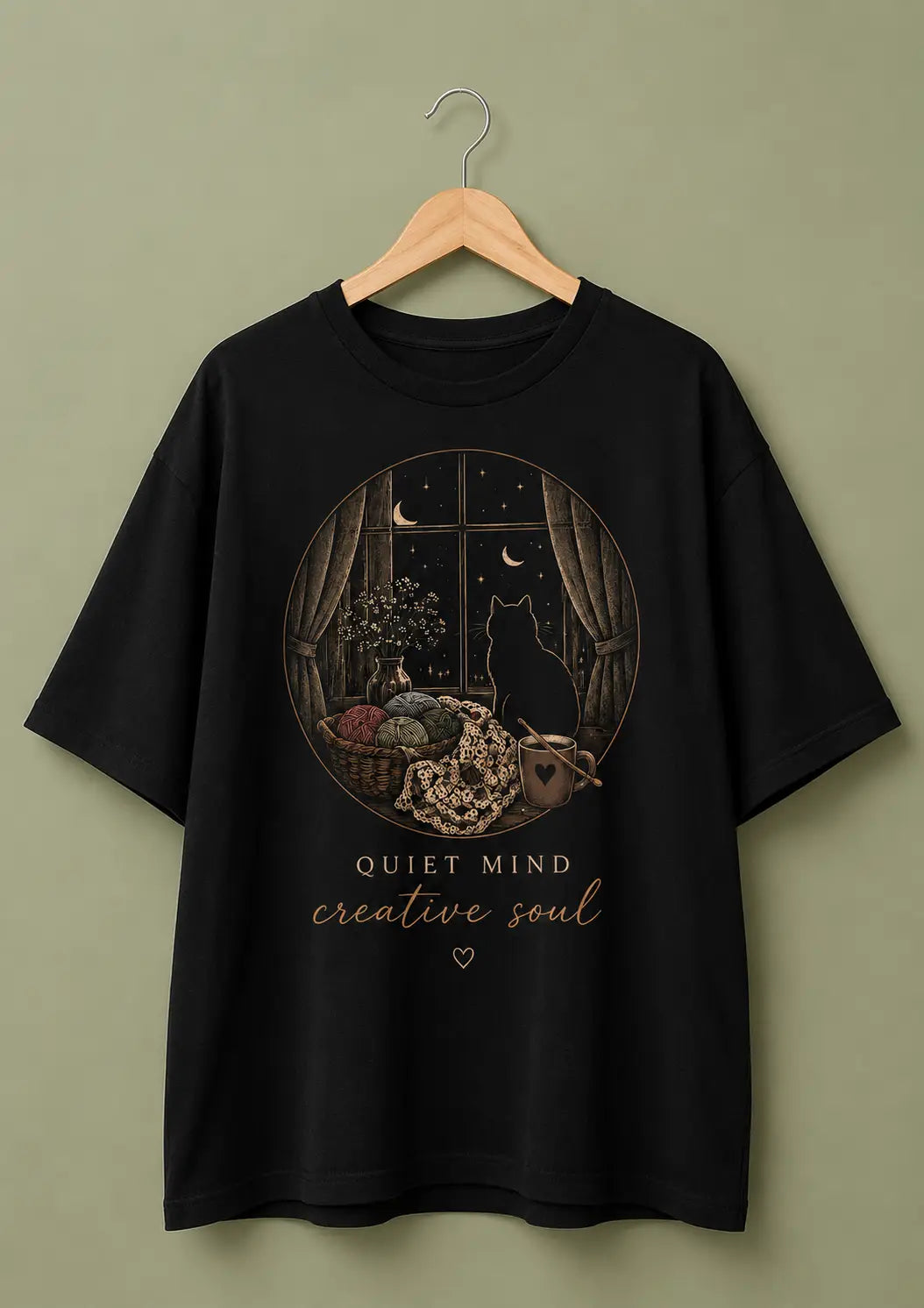 A black short-sleeve graphic t-shirt for crochet artists hanging on a wooden hanger against a muted olive-green background. The shirt features a circular graphic print of a black cat sitting by a window at night under a crescent moon, with a basket of colorful yarn, and the text 'QUIET MIND creative soul' printed below.