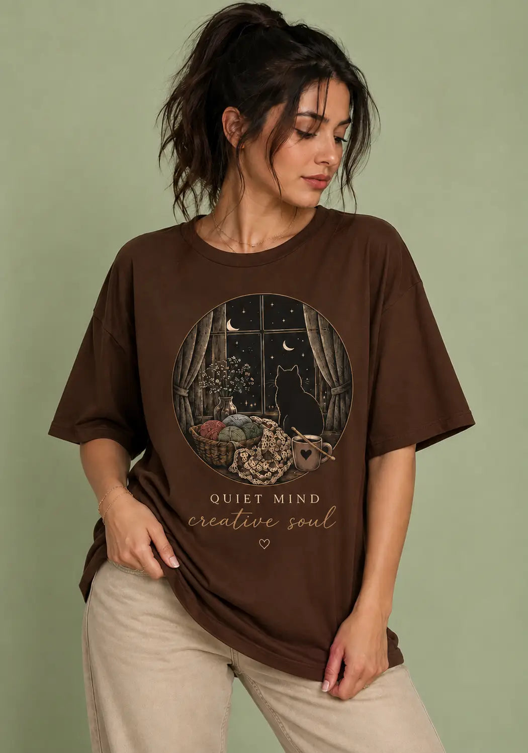 A woman wearing a brown graphic t-shirt for crochet artists with a circular illustration of a black cat in a room at night and the text 'QUIET MIND creative soul', paired with beige pants, set against a solid sage green background.