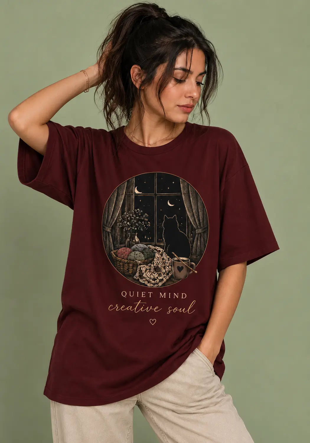 A woman wearing an oversized maroon graphic t-shirt for crochet artists with a circular illustration of a cat in a room and the text 'QUIET MIND creative soul', paired with light beige pants, set against a solid sage green background.