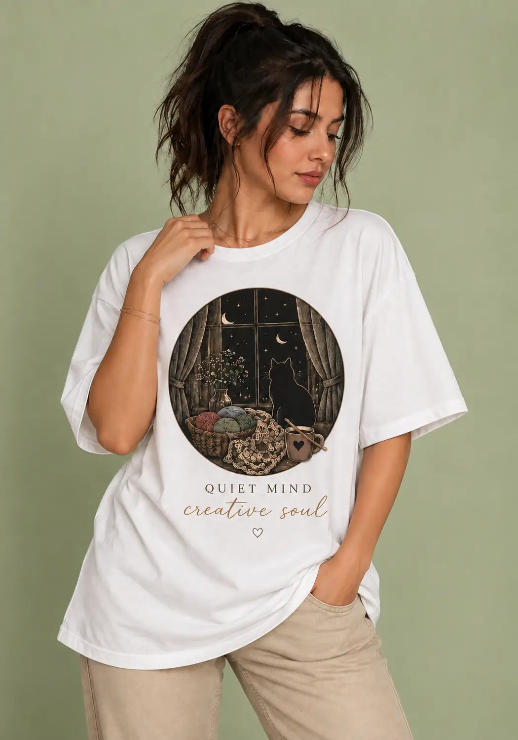 A woman wearing an oversized white graphic t-shirt for crochet artists with a circular illustration of a black cat in a room at night and the text 'QUIET MIND creative soul'. She is wearing beige pants against a solid sage green background.