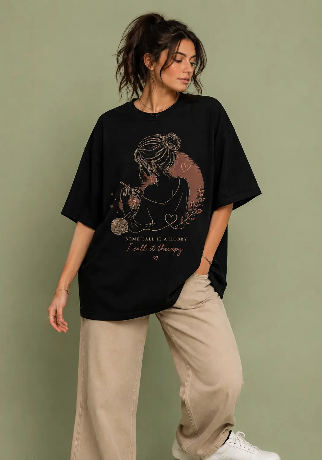 A woman modeling an oversized black graphic t-shirt for crochet artists with a rose gold illustration and text that reads 'Some call it a hobby I call it therapy', paired with beige wide-leg pants and white sneakers, set against a solid sage green background.