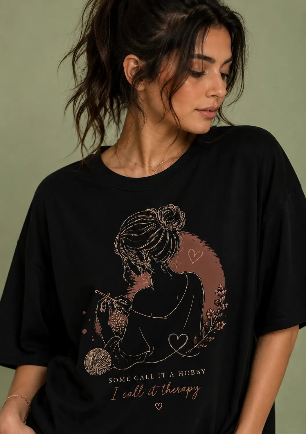 A woman wearing a black oversized graphic t-shirt for crochet artists with a rose gold line art illustration of a woman holding a mug, surrounded by hearts and the text 'SOME CALL IT A HONORRY I call it therapy'. The background is a solid sage green wall.