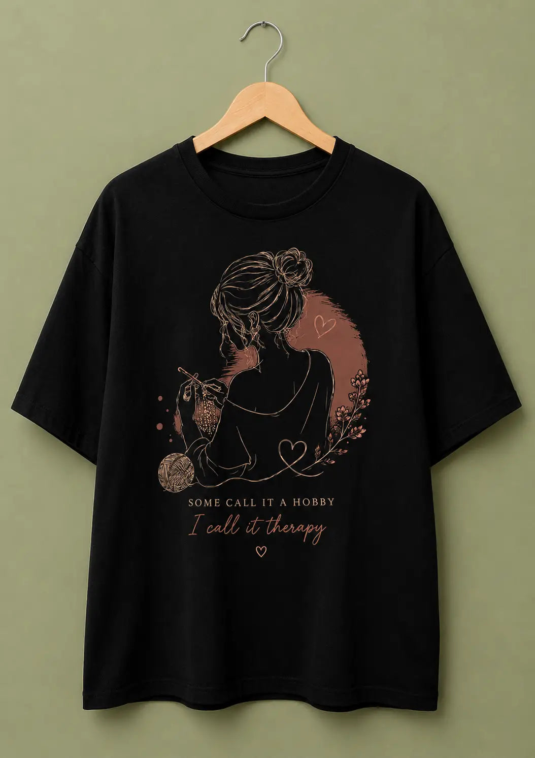A black short-sleeve t-shirt for crochet artists hanging on a wooden hanger against a solid olive-green background. The shirt features a graphic print of a woman in profile with a heart-shaped hair accessory, surrounded by small hearts and floral details, with the text 'SOME CALL IT A HOBBY, I call it therapy' printed below in a dusty rose color.
