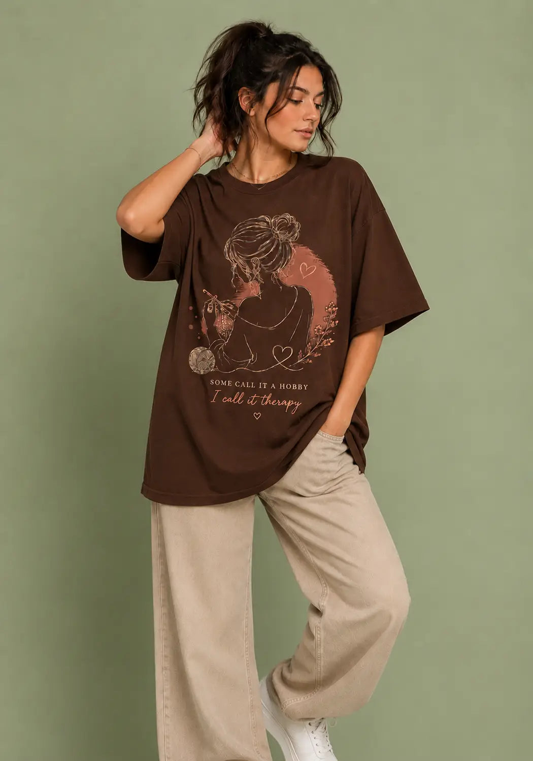 A woman modeling an oversized brown graphic t-shirt for crochet artists with a vintage-style illustration and the text 'YOU CAN'T HIDE A STORY, I tell it there', paired with beige wide-leg pants, against a solid sage green background.