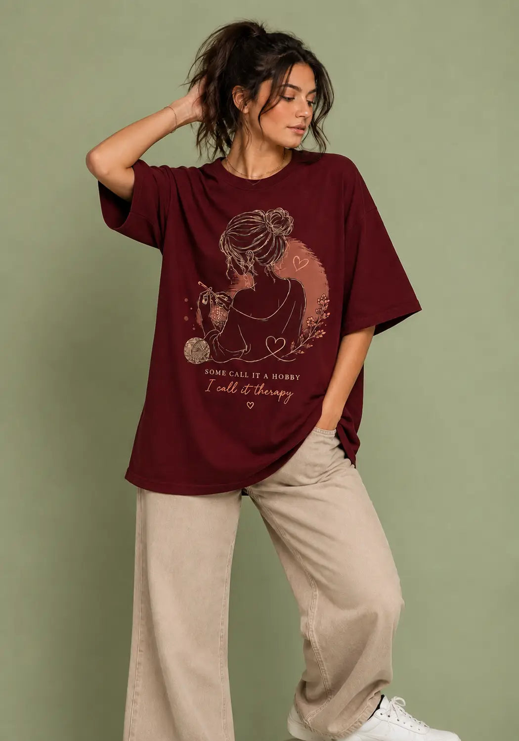 A woman modeling an oversized maroon graphic t-shirt for crochet artists with a cat illustration and the text 'YOUR CALL IS A HUNTER, I call it therapy', paired with beige wide-leg pants and white sneakers, set against a solid sage green background.