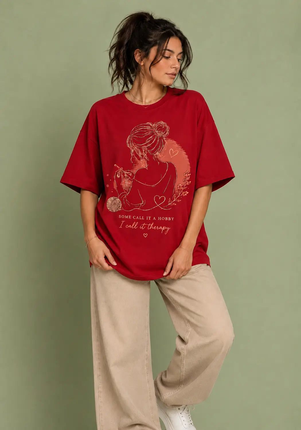 A woman modeling an oversized red graphic t-shirt for crochet artists with a pink line-art illustration of a woman and cat, paired with beige wide-leg pants, against a solid sage green background.