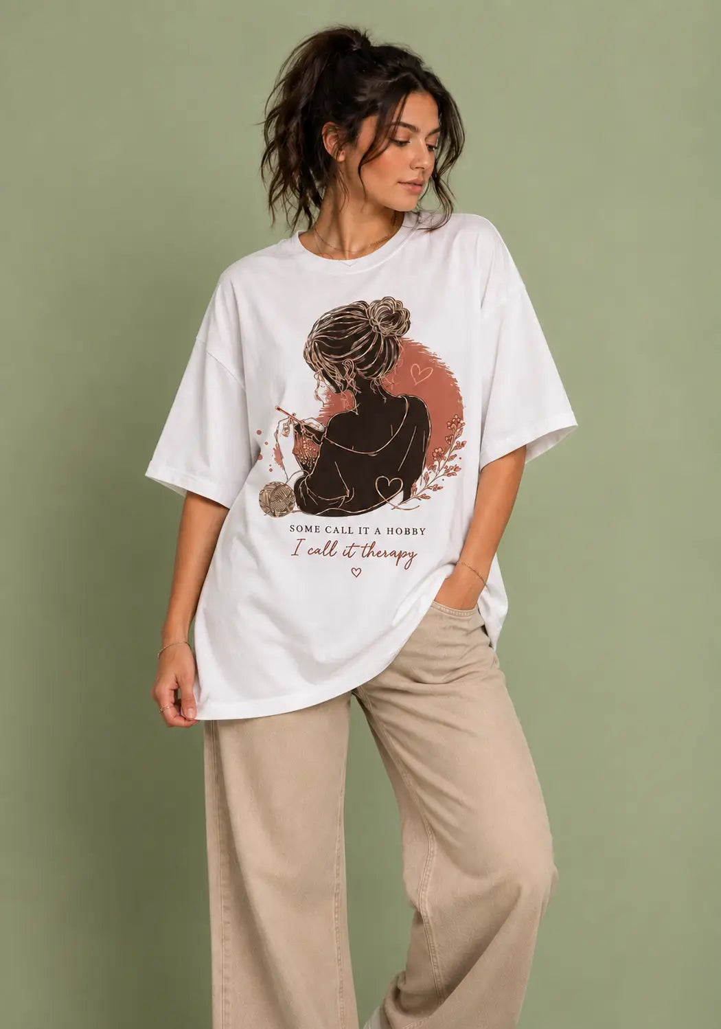 A woman wearing an oversized white graphic t-shirt for crochet artists with a brown and orange illustration of a woman holding a paintbrush, with the text 'I call it therapy' printed below. She is also wearing beige wide-leg pants, standing against a solid sage green background.