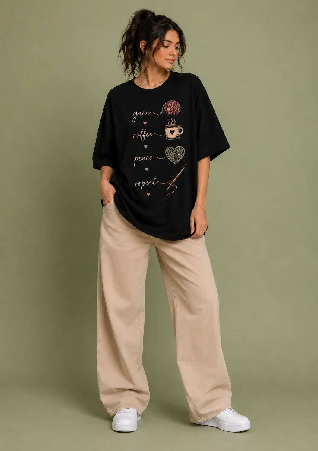 A woman modeling an oversized black graphic t-shirt for crochet artists with 'yarn coffee peace repeat' and small coffee cup/heart illustrations, paired with beige wide-leg pants and white sneakers, against a solid sage green studio background.