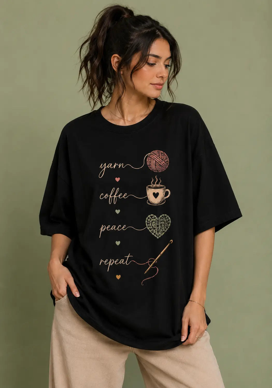 A woman wearing a black oversized graphic t-shirt for crochet artists with yarn, coffee, peace, and repeat designs, paired with beige pants, set against a solid olive-green background.