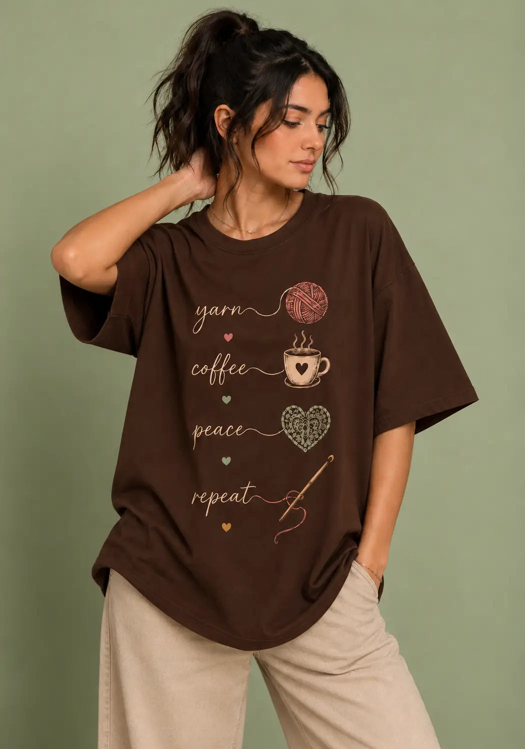 A woman wearing an oversized dark brown graphic t-shirt for crochet artists with yarn, coffee, peace, and repeat text and illustrations, paired with beige pants, set against a solid sage green background.