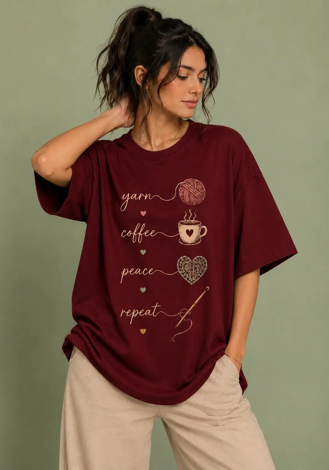 A woman wearing an oversized maroon graphic t-shirt for crochet artists with yarn, coffee, peace, and repeat text and illustrations, paired with beige pants, set against a solid sage green background.