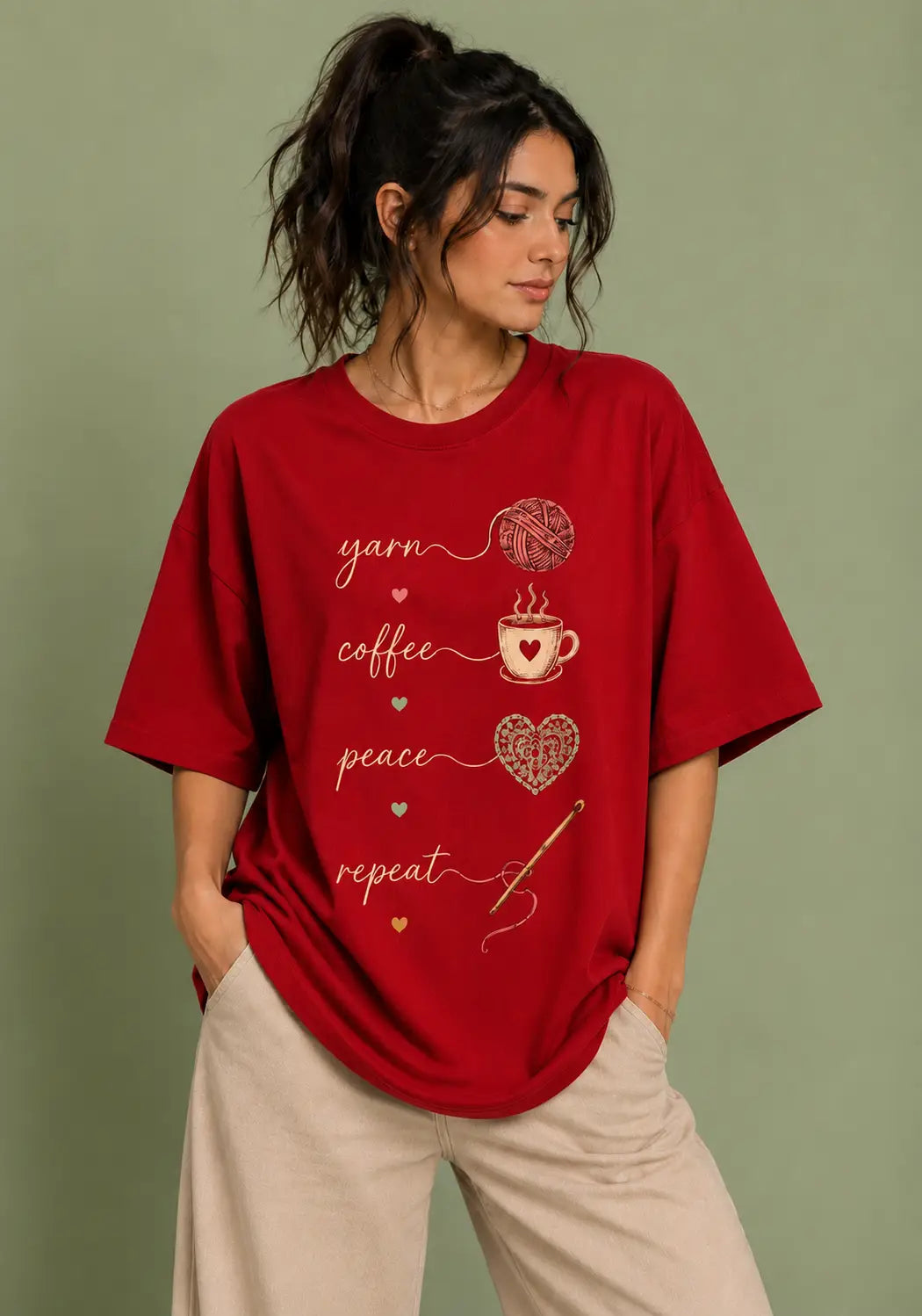 A woman wearing a red oversized t-shirt with for crochet artists a graphic print that reads 'yarn coffee peace repeat' in gold script with small heart and yarn motifs, paired with beige pants, set against a solid sage green background.