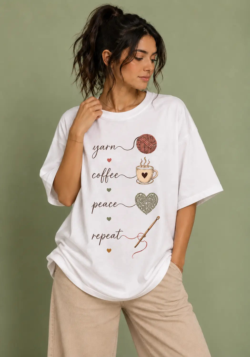 A woman wearing a white graphic t-shirt for crochet artists with yarn, coffee, peace, and repeat designs, paired with beige pants, set against a solid sage green background.