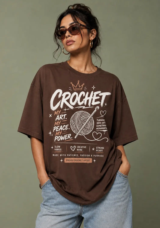 A woman wearing an oversized brown graphic t-shirt for crochet artists with crochet-themed text and illustrations, paired with light blue jeans, set against a solid olive-green background.