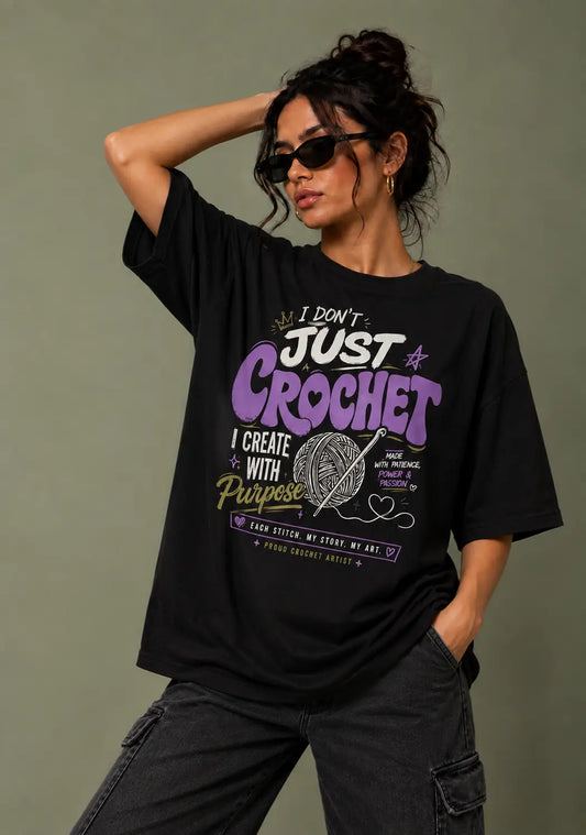 A woman wearing a black graphic t-shirt for crochet lover with purple and white text that reads 'I DON'T JUST CROCHET I CREATE WITH PURPOSE' and a yarn ball illustration, paired with dark gray cargo pants against a solid olive-gray background.