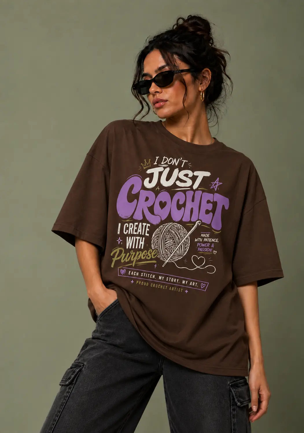 A woman wearing an oversized brown graphic t-shirt for crochet lover with purple and white text that reads 'I DON'T JUST CROCHET I CREATE WITH PURPOSE' and a yarn ball illustration, paired with dark gray cargo pants against a muted olive background.
