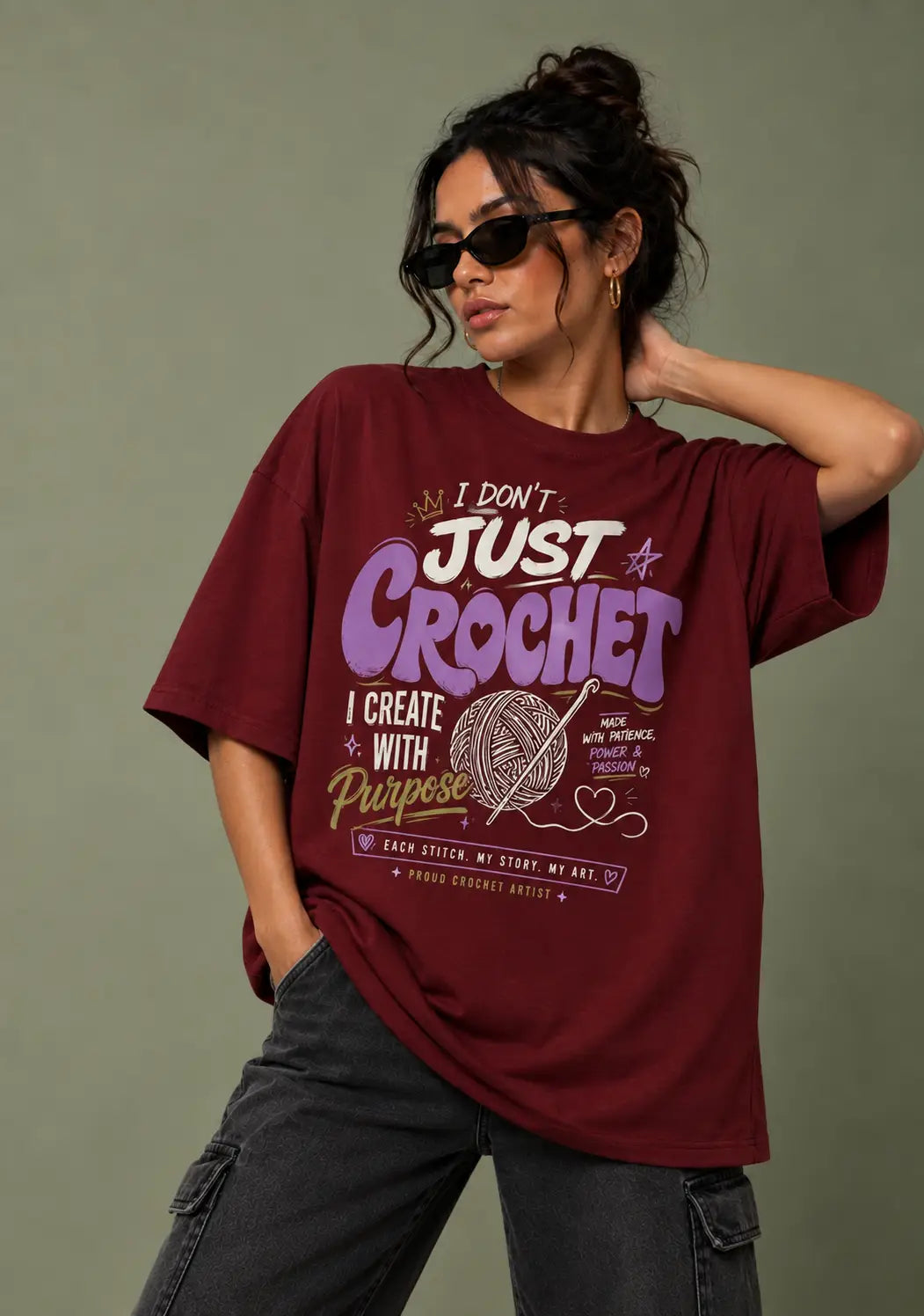A woman wearing a maroon graphic t-shirt for crochet lover with a crochet-themed print that reads 'I DON'T JUST CROCHET I CREATE WITH Purpose' and 'BROOK STITCH, MY STORY, MY ART.' She is also wearing dark sunglasses and dark gray cargo pants, posed against a solid olive-green background.