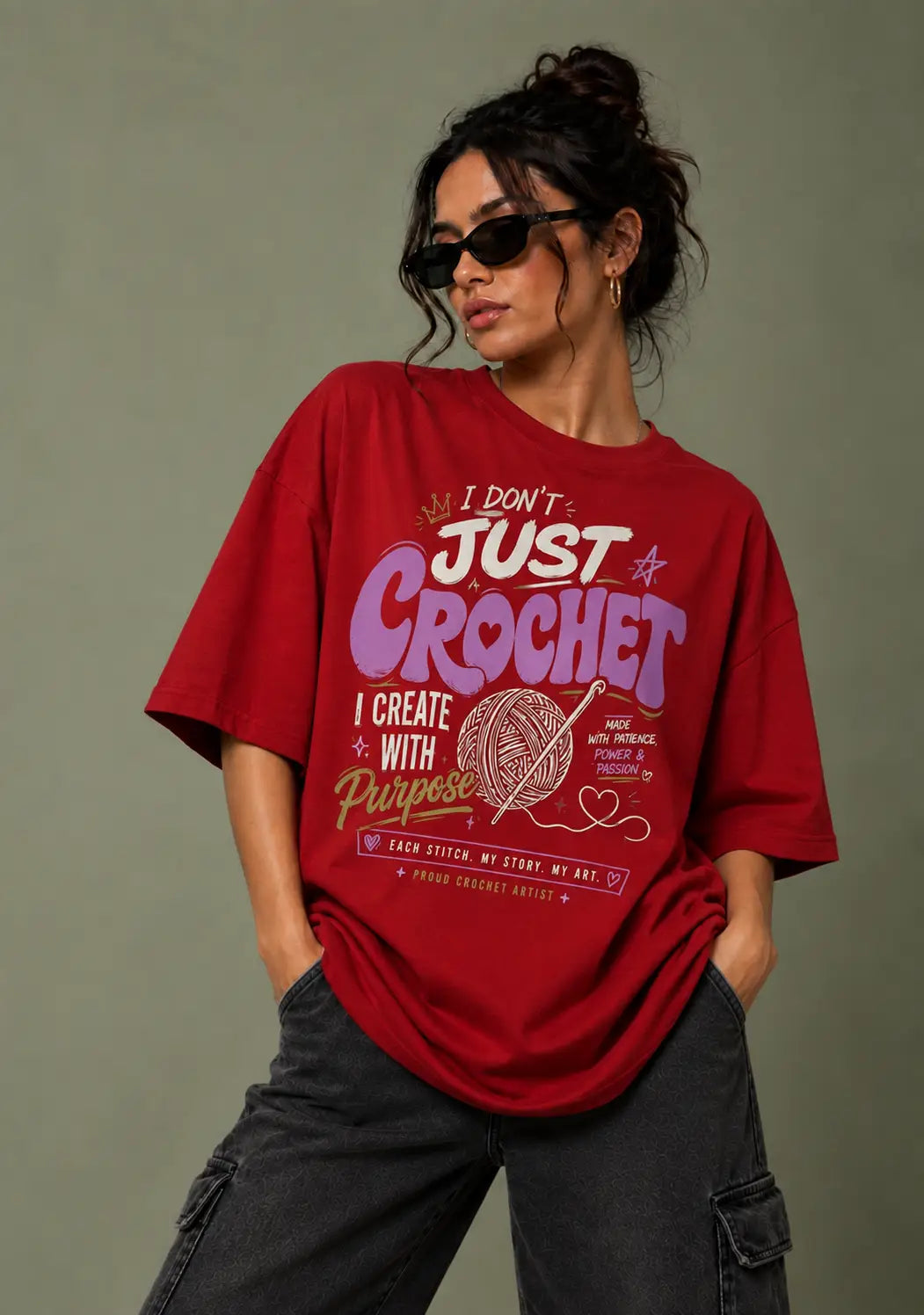A woman wearing a red graphic t-shirt for crochet lover with a crochet-themed print that reads 'I DON'T JUST CROCHET I CREATE WITH Passion'. She is wearing black cargo pants and sunglasses, posing against a solid gray background.
