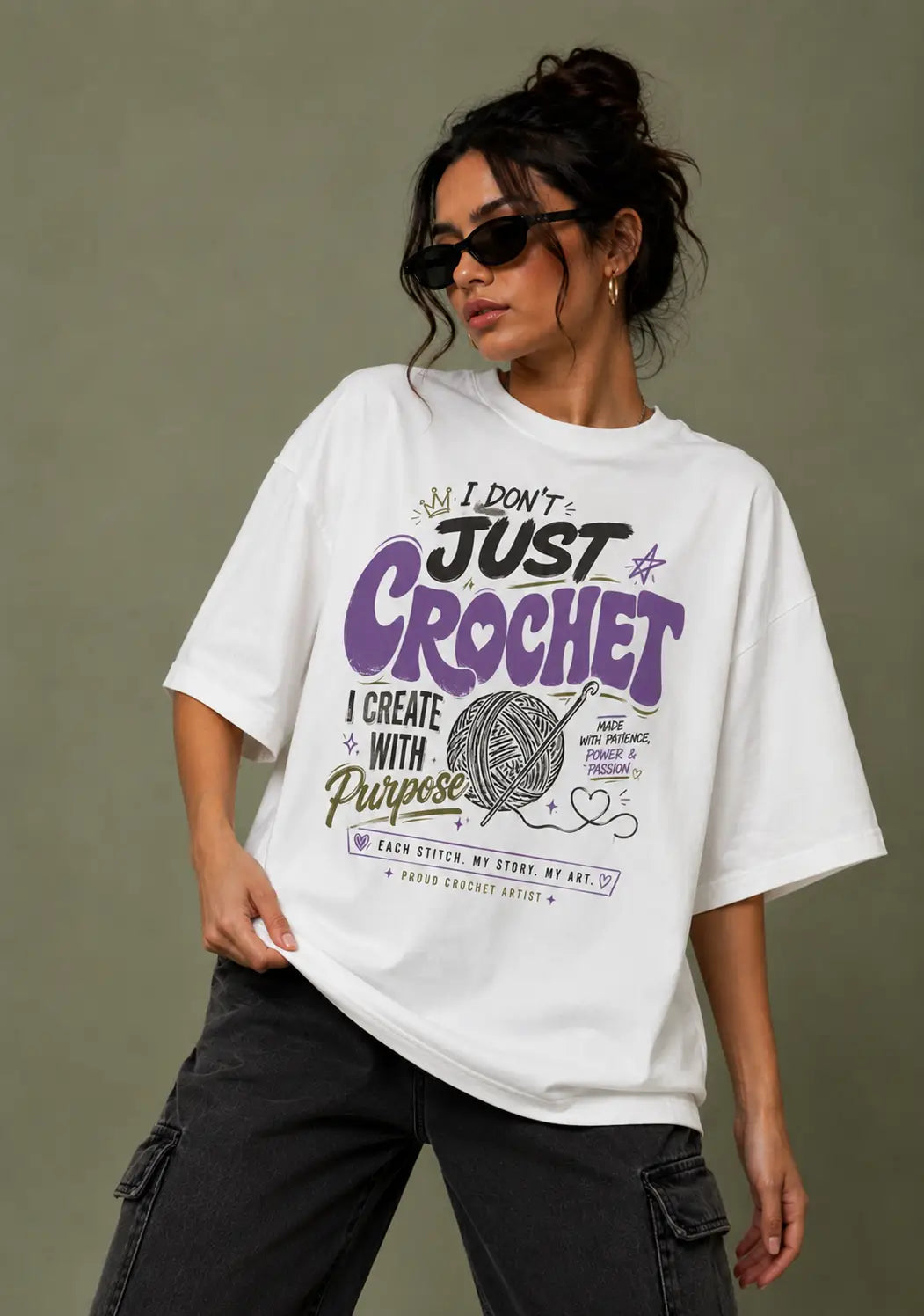 A woman wearing a white oversized graphic T-shirt for crochet lover with purple and black text that reads 'I DON'T JUST CROCHET I CREATE WITH Purpose' along with a yarn ball illustration, paired with dark cargo pants against a muted olive-gray background.