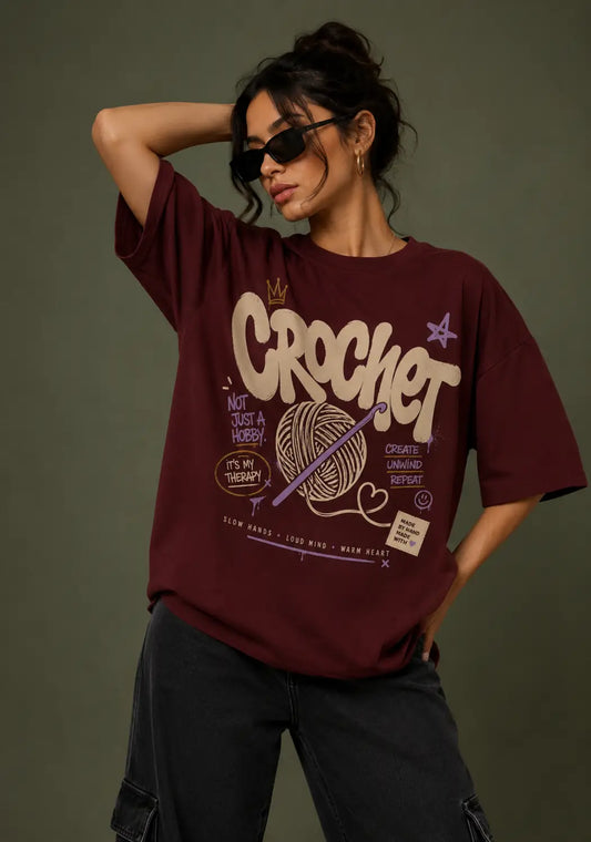 A woman wearing a maroon oversized graphic T-shirt for crochet artists with crochet-themed text and yarn ball illustration, paired with dark cargo pants, posed against a dark olive background.