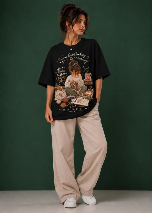 A woman stands against a dark green studio backdrop wearing an oversized black graphic T-shirt for crochet artists with a vintage-style illustration and text, loose light beige pants, and white sneakers.