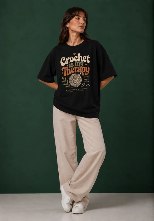 A woman stands against a dark green studio backdrop wearing a black crochet artists graphic T-shirt with the phrase 'Crochet is my Therapy' and a yarn ball illustration, paired with light beige wide-leg pants and white sneakers.