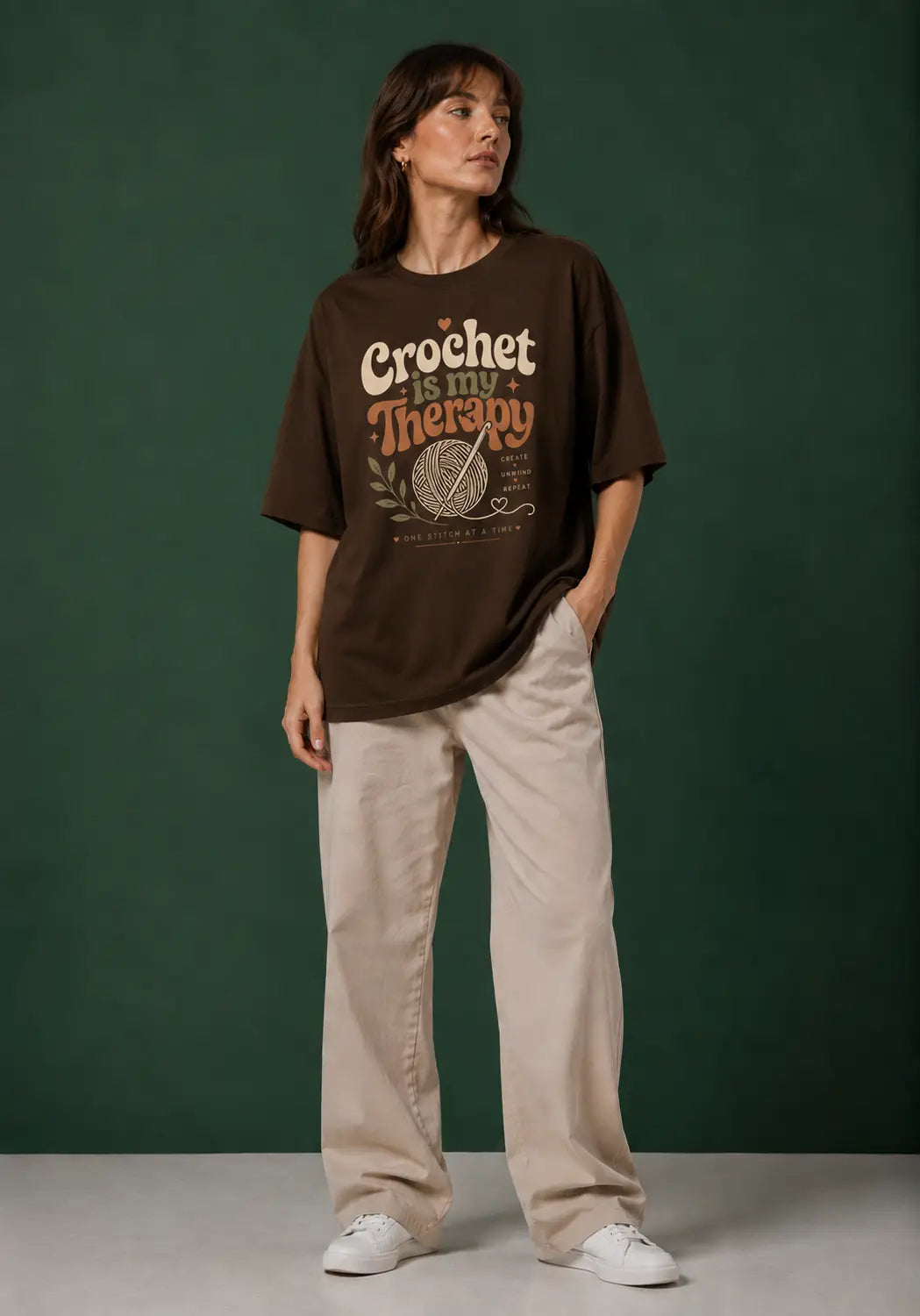 A woman stands against a dark green studio backdrop wearing an oversized brown crochet artist's graphic T-shirt with the words 'Crochet is my Therapy' and light beige wide-leg pants with white sneakers.
