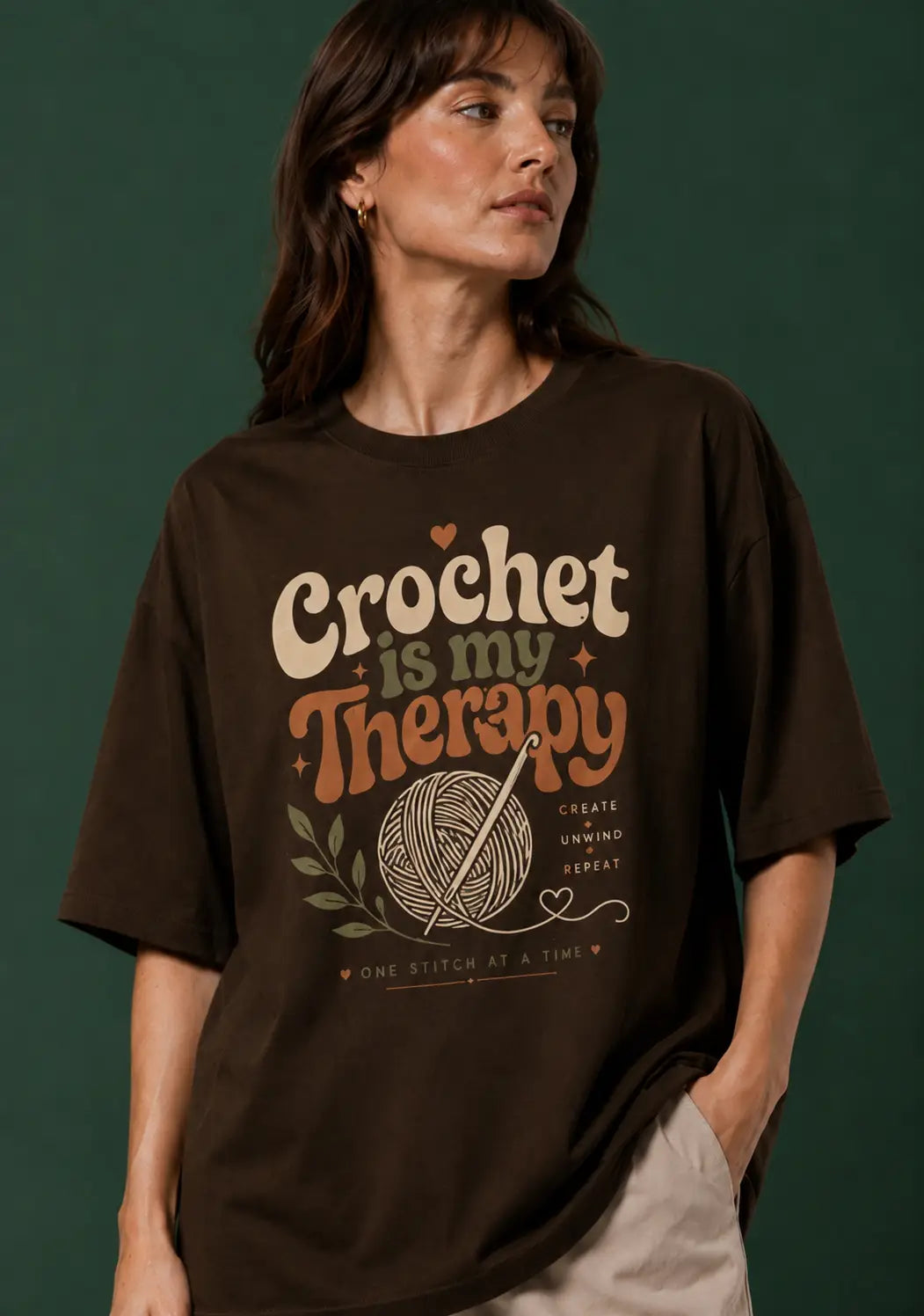 A woman wearing an oversized dark brown graphic T-shirt for crochet artists with the text 'Crochet is my Therapy' and a crochet ball illustration, posed against a dark green background.
