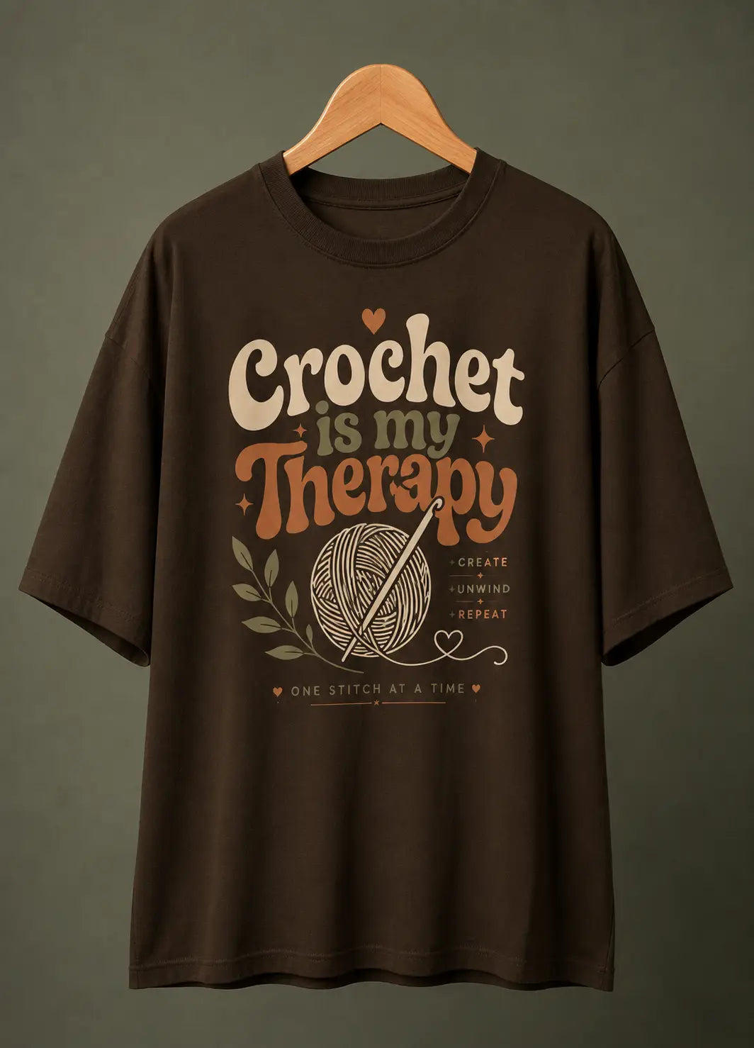 A dark brown short-sleeve t-shirt hanging on a wooden hanger against a dark gray background. The shirt features a retro-style graphic print that reads 'Crochet is my Therapy' in cream and orange lettering, accompanied by a yarn ball and crochet hook illustration.
