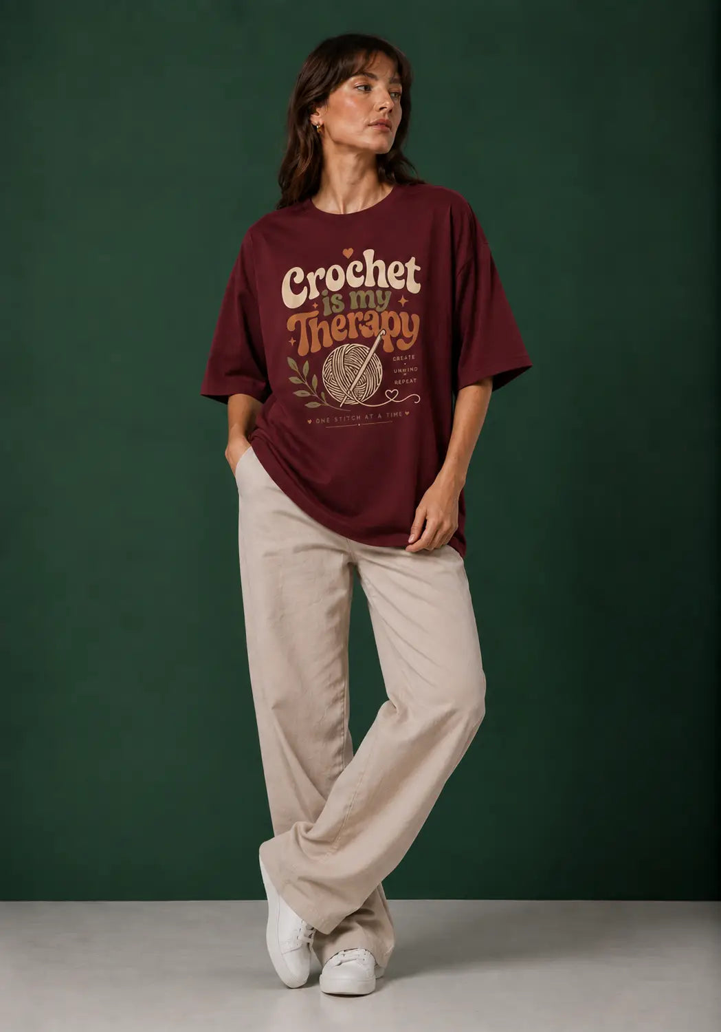 A woman stands against a dark green studio backdrop wearing an oversized burgundy graphic T-shirt with for crochet artists cream and orange text reading 'Crochet is my Therapy,' paired with light beige wide-leg pants and white sneakers.