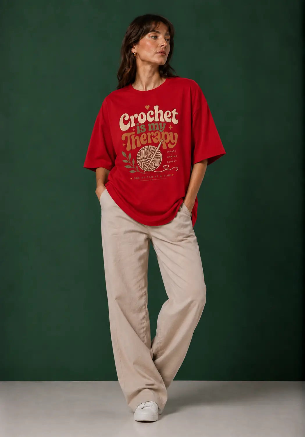 A woman modeling an oversized red graphic T-shirt  for crochet artists with cream text reading 'Crochet is my Therapy' and a yarn ball illustration, paired with light beige wide-leg pants and white sneakers, against a dark green studio backdrop.