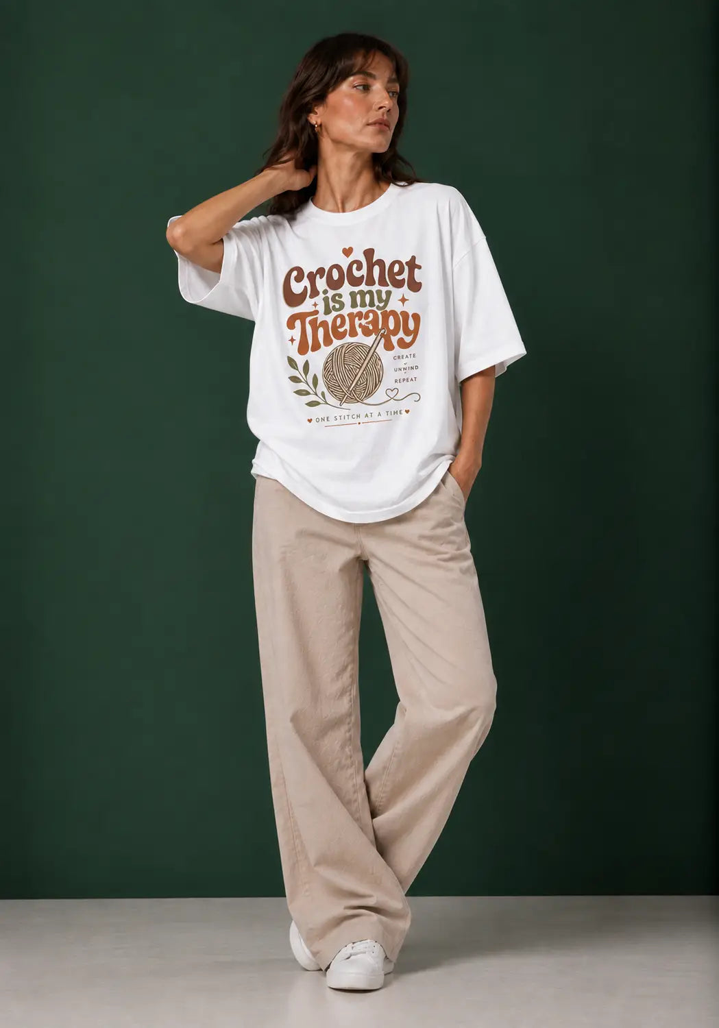 A woman modeling a white graphic t-shirt for crochet artists with the text 'Crochet is my Therapy' in brown and orange lettering, paired with beige wide-leg trousers, against a dark green studio backdrop.