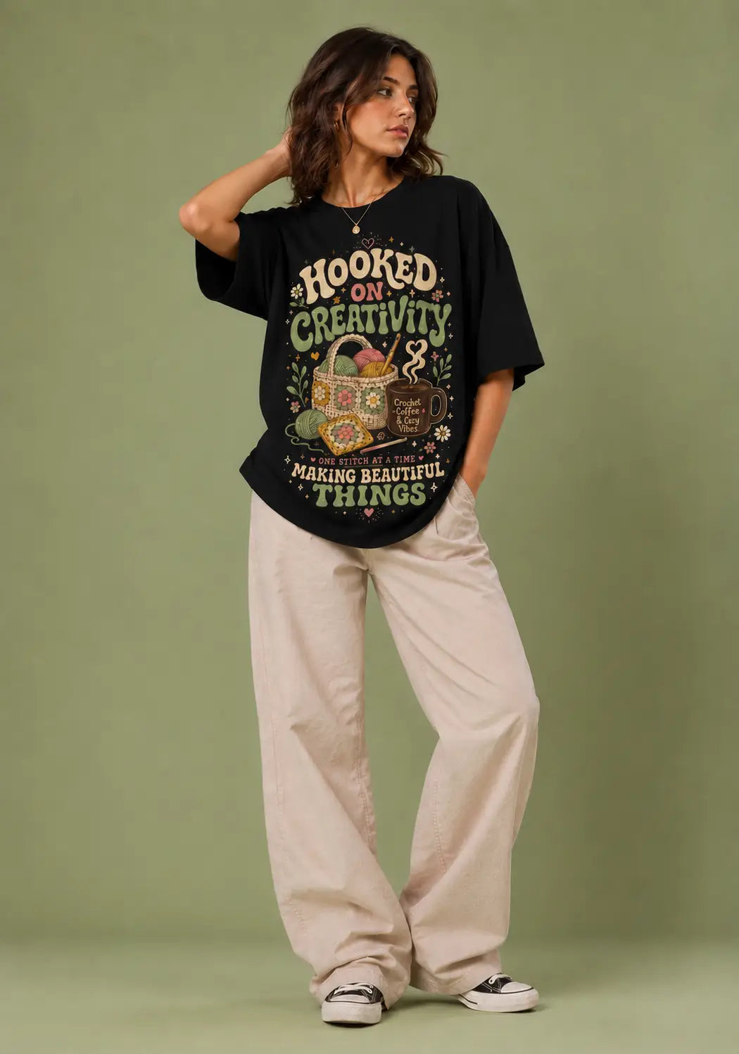 A woman modeling a black graphic t-shirt with a vintage-style design that reads 'HOOKED ON CREATIVITY! MAKING BEAUTIFUL THINGS!' in cream and green, paired with beige wide-leg pants and black-and-white sneakers, set against a solid sage green studio backdrop.
