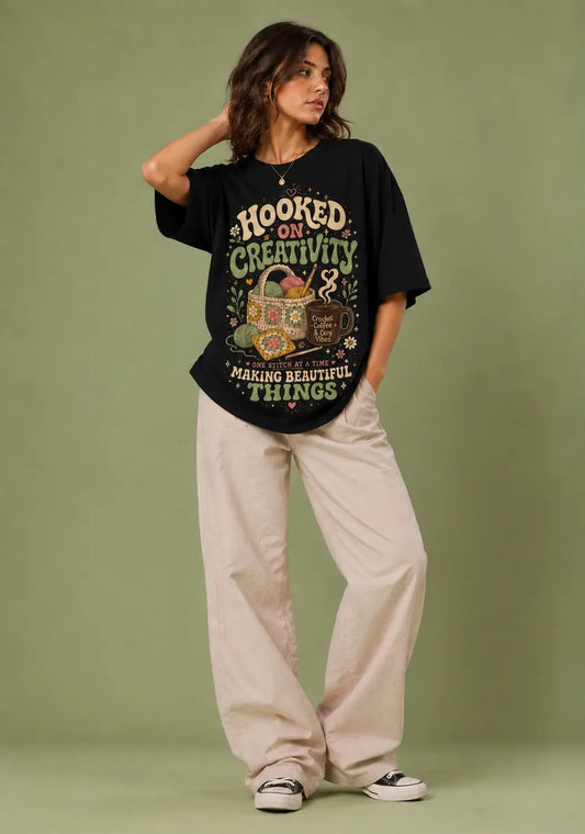 A woman modeling a black graphic t-shirt with a vintage-style design that reads 'HOOKED ON CREATIVITY! MAKING BEAUTIFUL THINGS!' in cream and green, paired with beige wide-leg pants and black-and-white sneakers, set against a solid sage green studio backdrop.