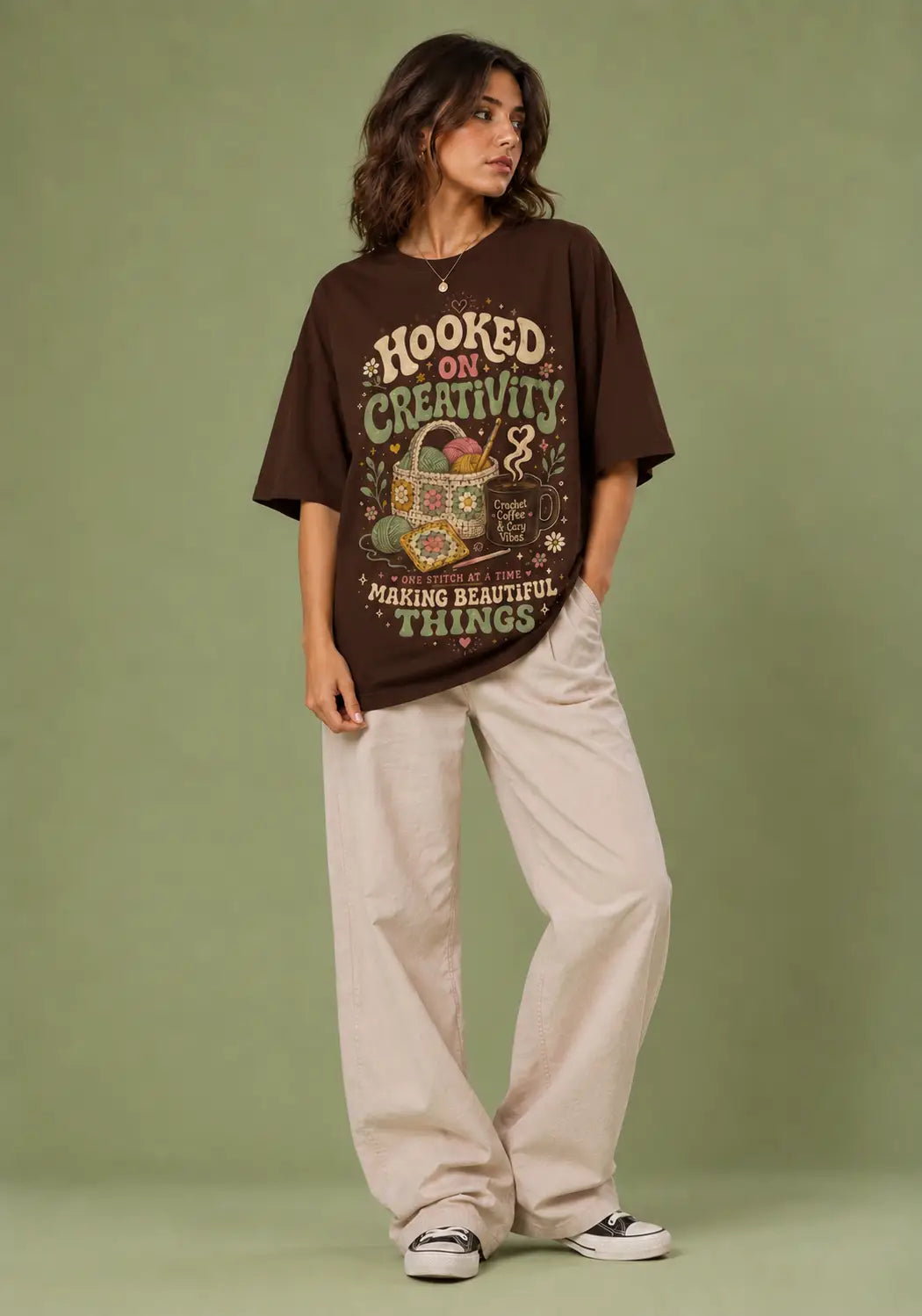 A woman modeling an oversized dark brown graphic t-shirt with retro-style text and illustration, paired with light beige wide-leg pants and black-and-white sneakers, set against a solid sage green studio background.