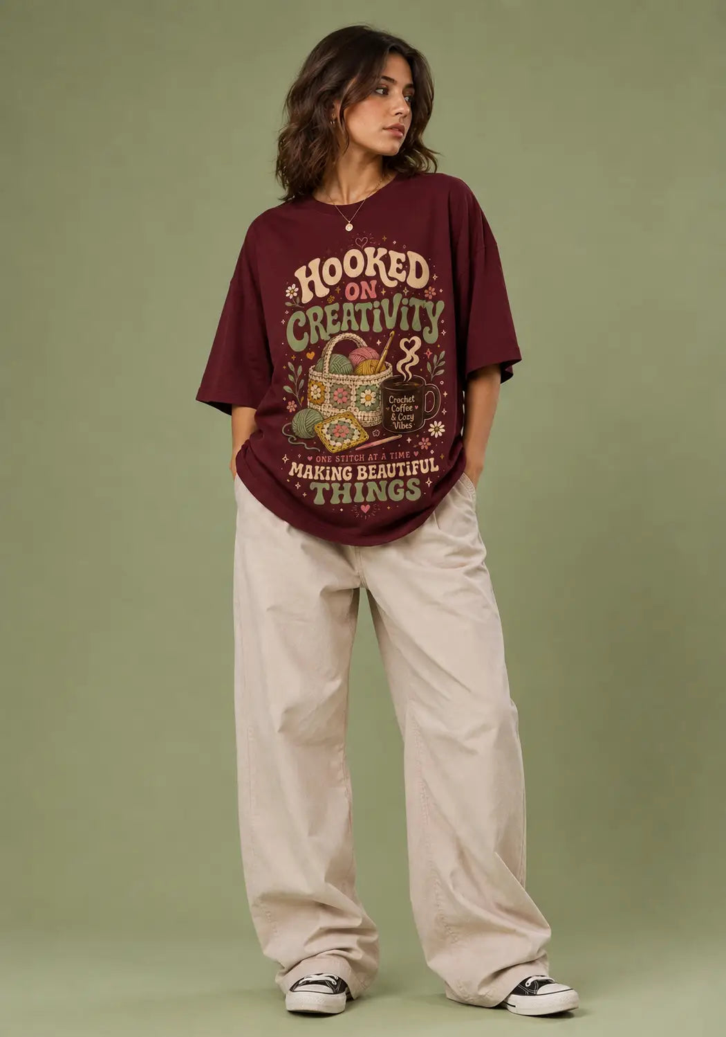 A woman modeling an oversized maroon graphic t-shirt with cream and green text reading 'HOOKED ON CREATIVITY MAKING BEAUTIFUL THINGS', paired with beige wide-leg pants and black-and-white sneakers, against a solid sage green studio background.