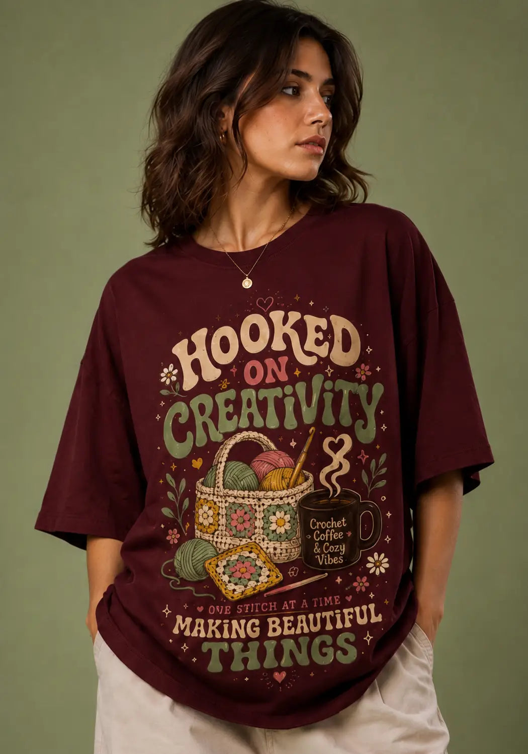 A woman wearing an oversized maroon graphic t-shirt with the text 'HOOKED ON CREATIVITY' and 'MAKING BEAUTIFUL THINGS' in a crochet-themed illustration, posed against a solid olive green background.