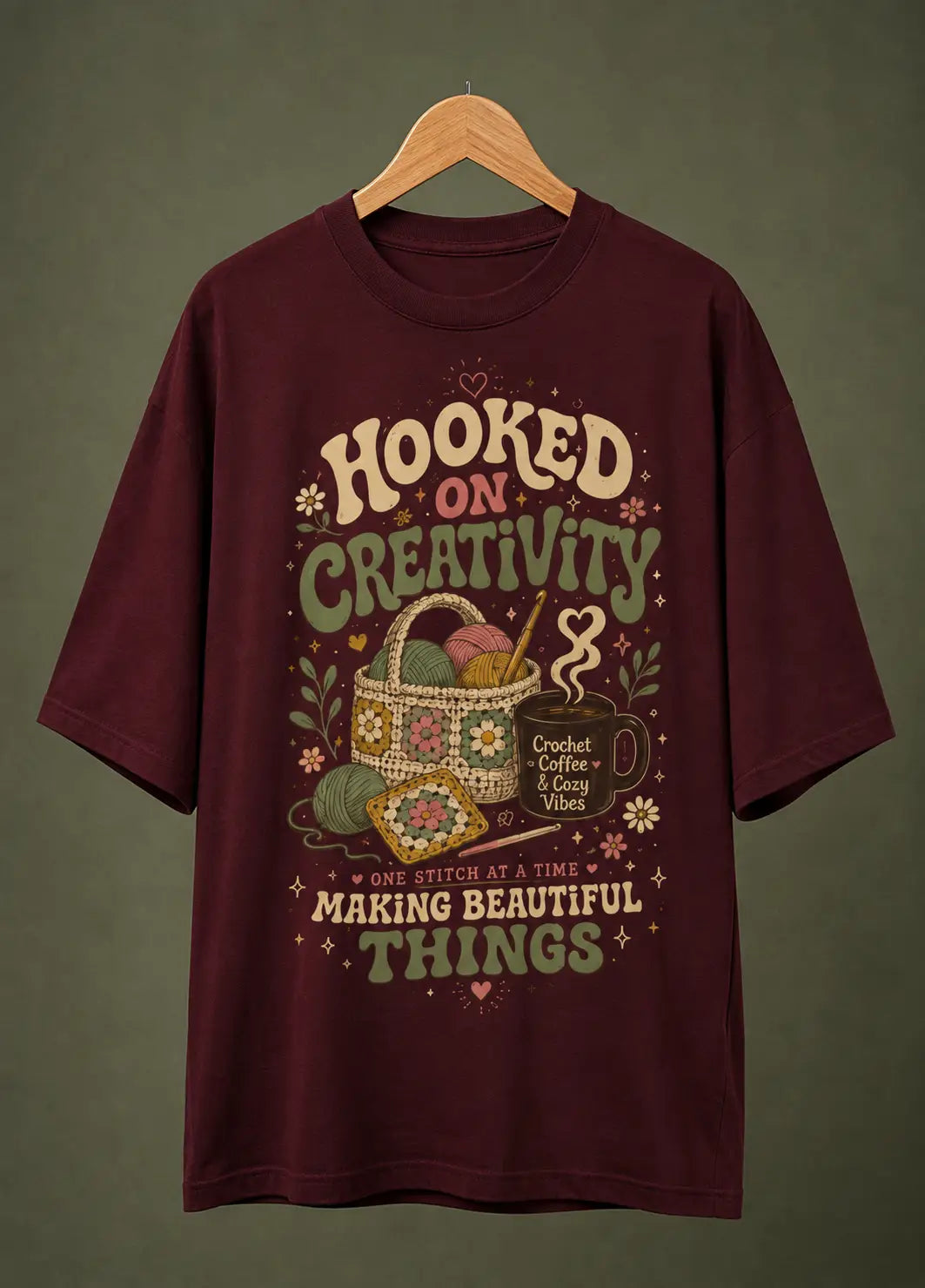 A maroon short-sleeve t-shirt hanging on a wooden hanger against a dark olive green background. The shirt features a retro-style graphic with the text 'HOOKED ON CREATIVITY' and 'MAKING BEAUTIFUL THINGS' surrounding an illustration of a crocheted basket, yarn balls, a mug, and crochet hooks.