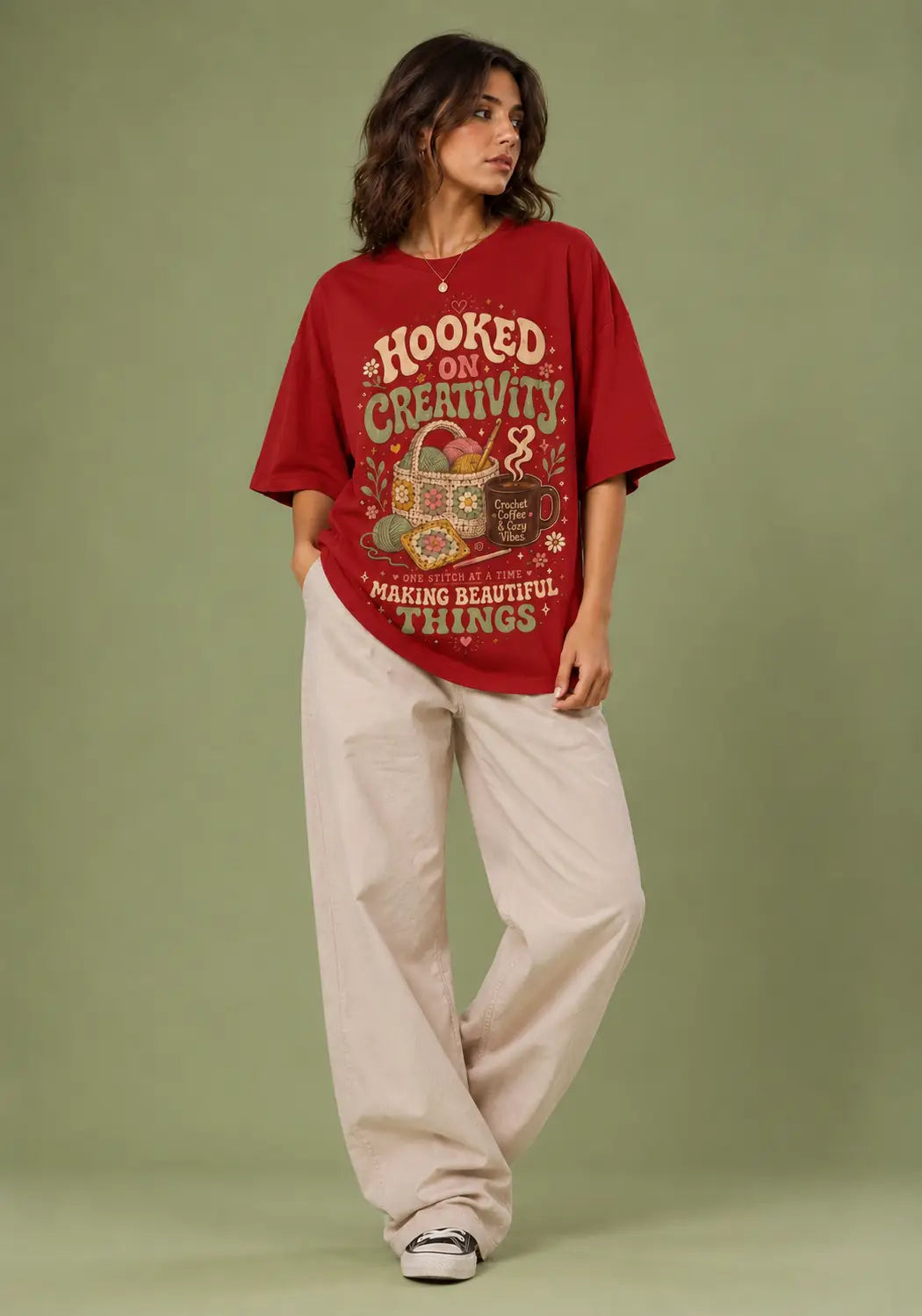 A woman modeling an oversized red graphic t-shirt with cream lettering that reads 'HOOKED ON CREATIVITY MAKING BEAUTIFUL THINGS', paired with light beige wide-leg pants, against a solid sage green studio background.