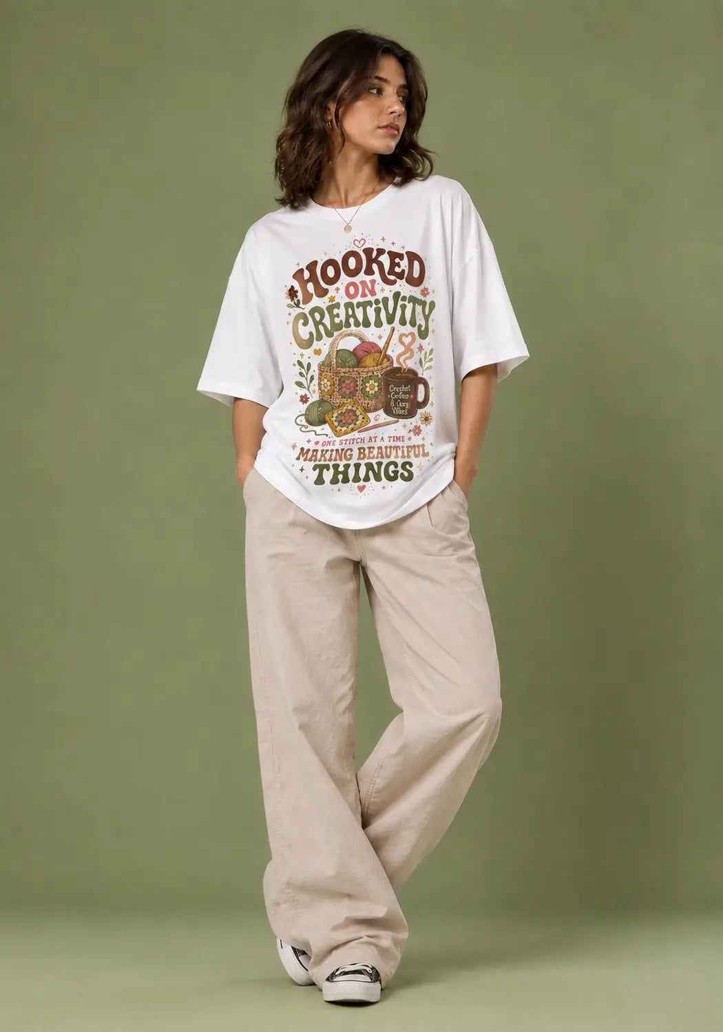 A woman modeling an oversized white graphic T-shirt with the text 'HOOKED ON CREATIVITY' and 'MINDING BEAUTIFUL THINGS' over a colorful illustration, paired with beige wide-leg pants against a solid sage green studio background.