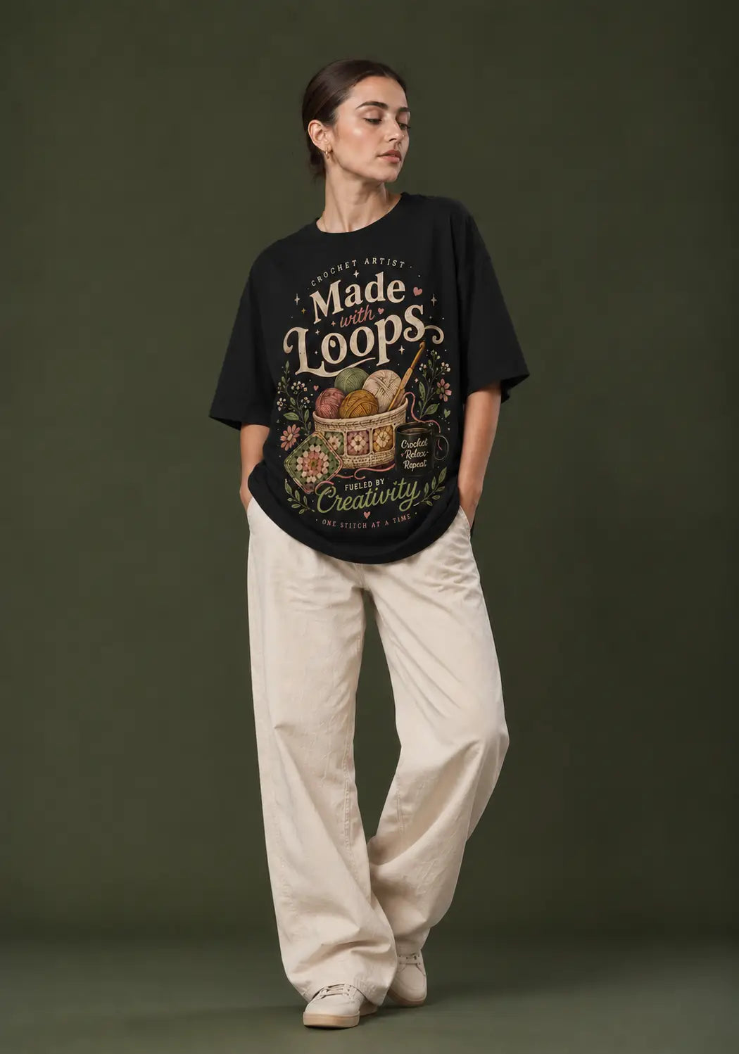 A woman modeling a black graphic t-shirt with a vintage-style crochet lover print that reads 'Made by Loops' and 'Creativity is a daily act', paired with cream-colored wide-leg pants, set against a dark olive green studio background.