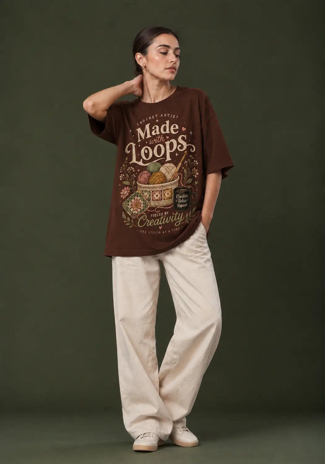 A woman stands against a dark olive green studio background wearing an oversized brown graphic crochet artist T-shirt with cream text reading 'Made of Loops' and a colorful illustration, paired with loose cream pants and white sneakers.