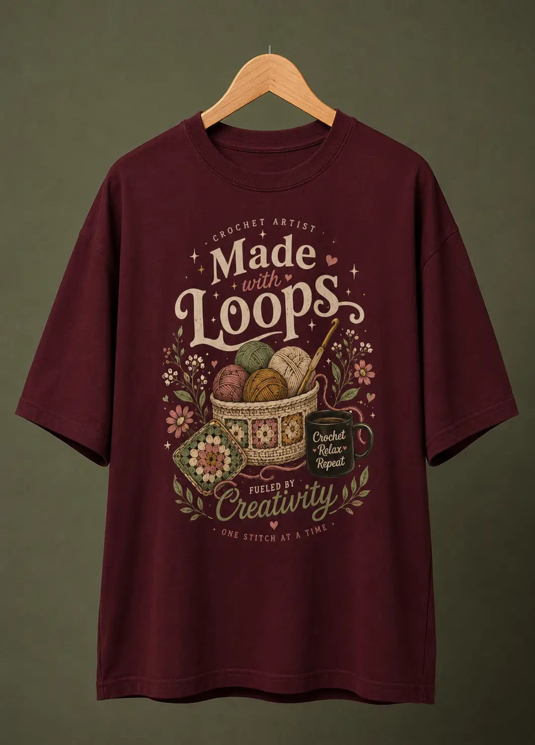 A maroon short-sleeve t-shirt hanging on a wooden hanger against a dark olive green background. The shirt features a detailed graphic print in the center with the text 'Made with Loops' and 'Crochet Artist' surrounded by crochet hooks, yarn balls, flowers, and a mug.