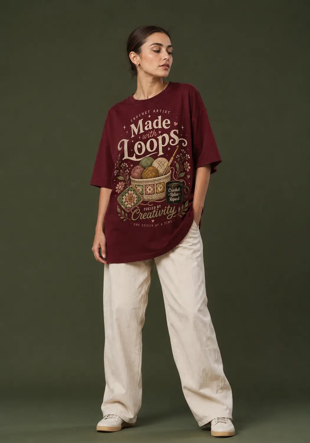 A woman modeling an oversized maroon crochet artist graphic T-shirt with cream text reading 'Made of Loops' and a vintage-style illustration, paired with off-white wide-leg pants and white sneakers, against a dark olive green studio background.