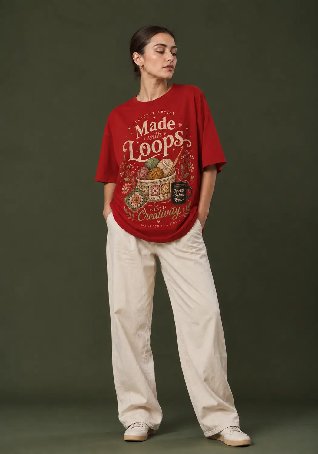 A woman stands against a dark olive green studio background wearing an oversized red graphic crochet lover T-shirt with cream lettering that reads 'Made from Loops' and cream wide-leg pants.