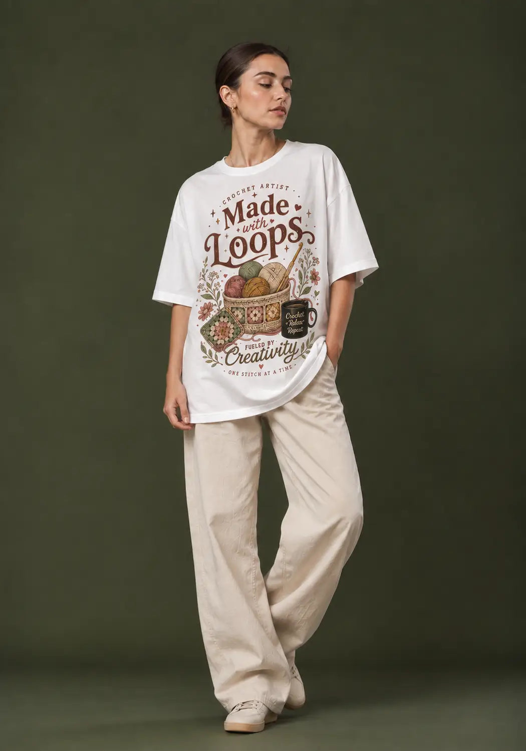 A woman stands against a dark olive green studio background wearing an oversized white crochet lover  graphic T-shirt and loose beige pants. The shirt features a vintage-style multicolor print with the words 'Made by the Loops' and 'Creativity'.