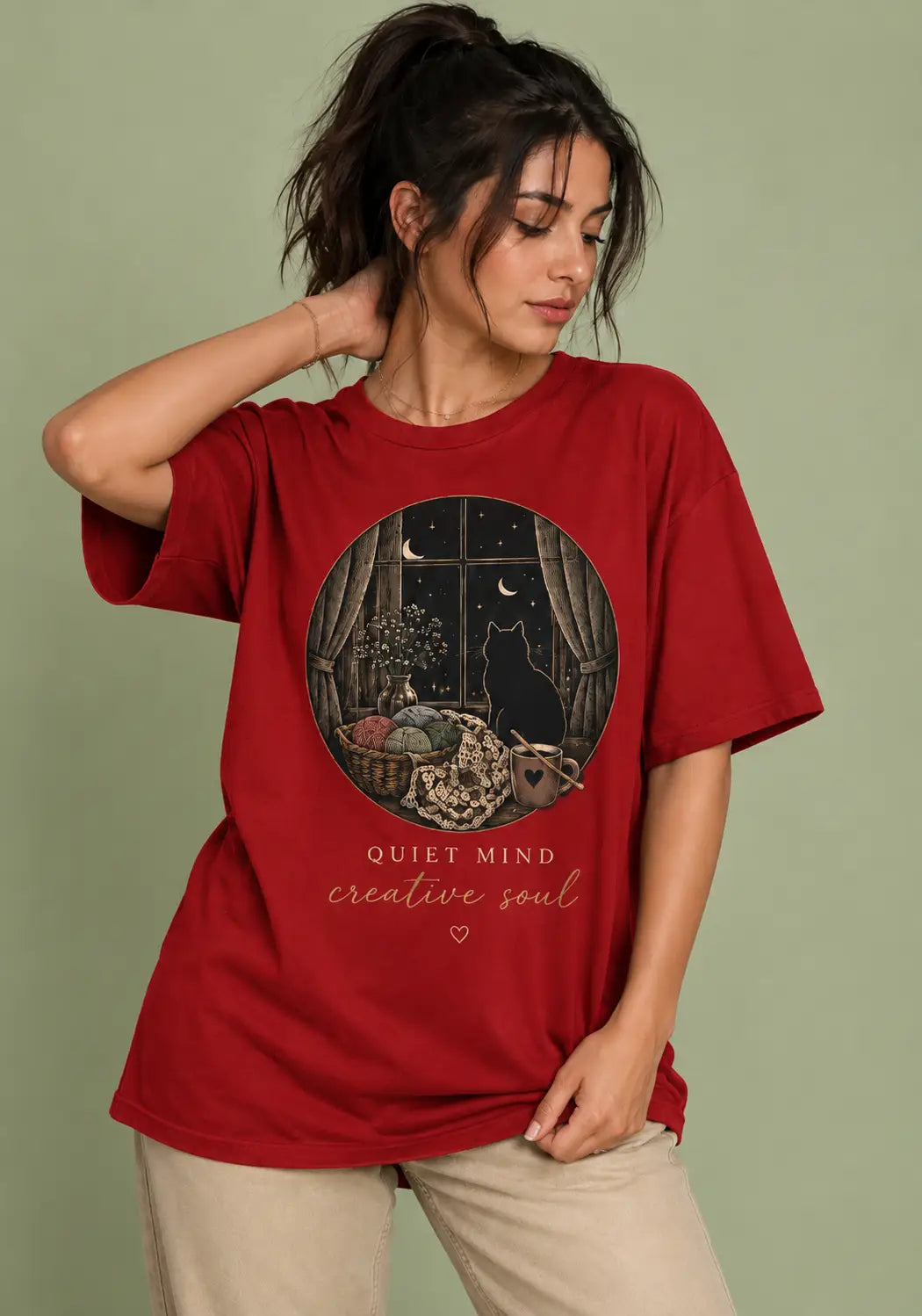 A woman wearing a red graphic t-shirt for crochet artists with a circular illustration of a cat in a window and the text 'QUIET MIND creative soul', paired with beige pants, set against a solid sage green background.